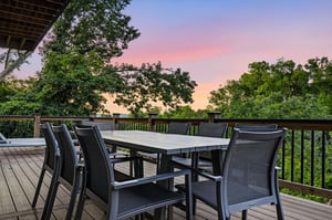 The propertys spacious deck provides a peaceful outdoor dining setting surrounded by mature trees and natural greenery, ideal for evening gatherings under colorful skies.