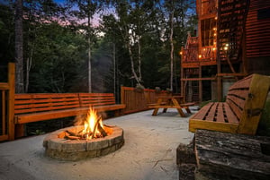 A rustic fire pit anchors this paved outdoor patio with wooden benches and picnic table, surrounded by forest. The cabins warm deck lights create a welcoming evening gathering spot.