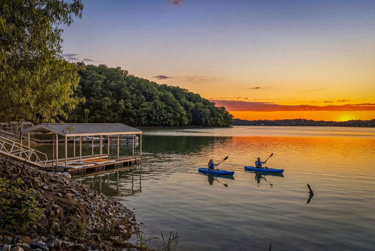 Sunset paints the sky over tranquil lake waters where kayakers glide peacefully, surrounded by wooded hills and a private dock at the propertys edge.