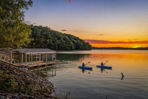 Sunset paints the sky over tranquil lake waters where kayakers glide peacefully, surrounded by wooded hills and a private dock at the propertys edge.