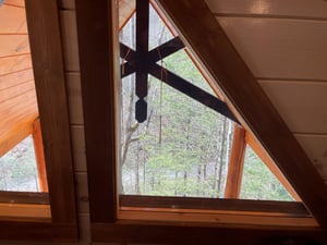 Relax in your cozy loft retreat with stunning forest views through charming triangular windows framed by warm wooden beams.