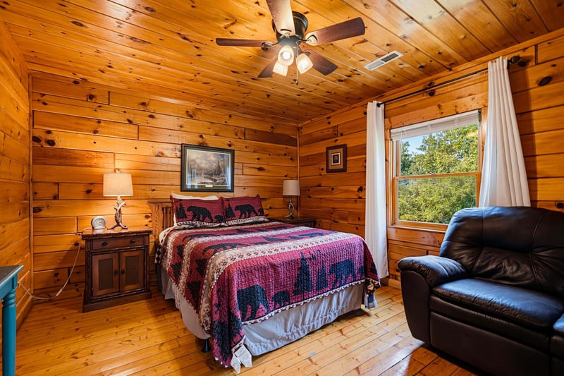Wake up in this warm, rustic bedroom where knotty pine walls and a soft quilted bed create your cozy cabin retreat. Natural light streams through the window, inviting you to start your day refreshed.