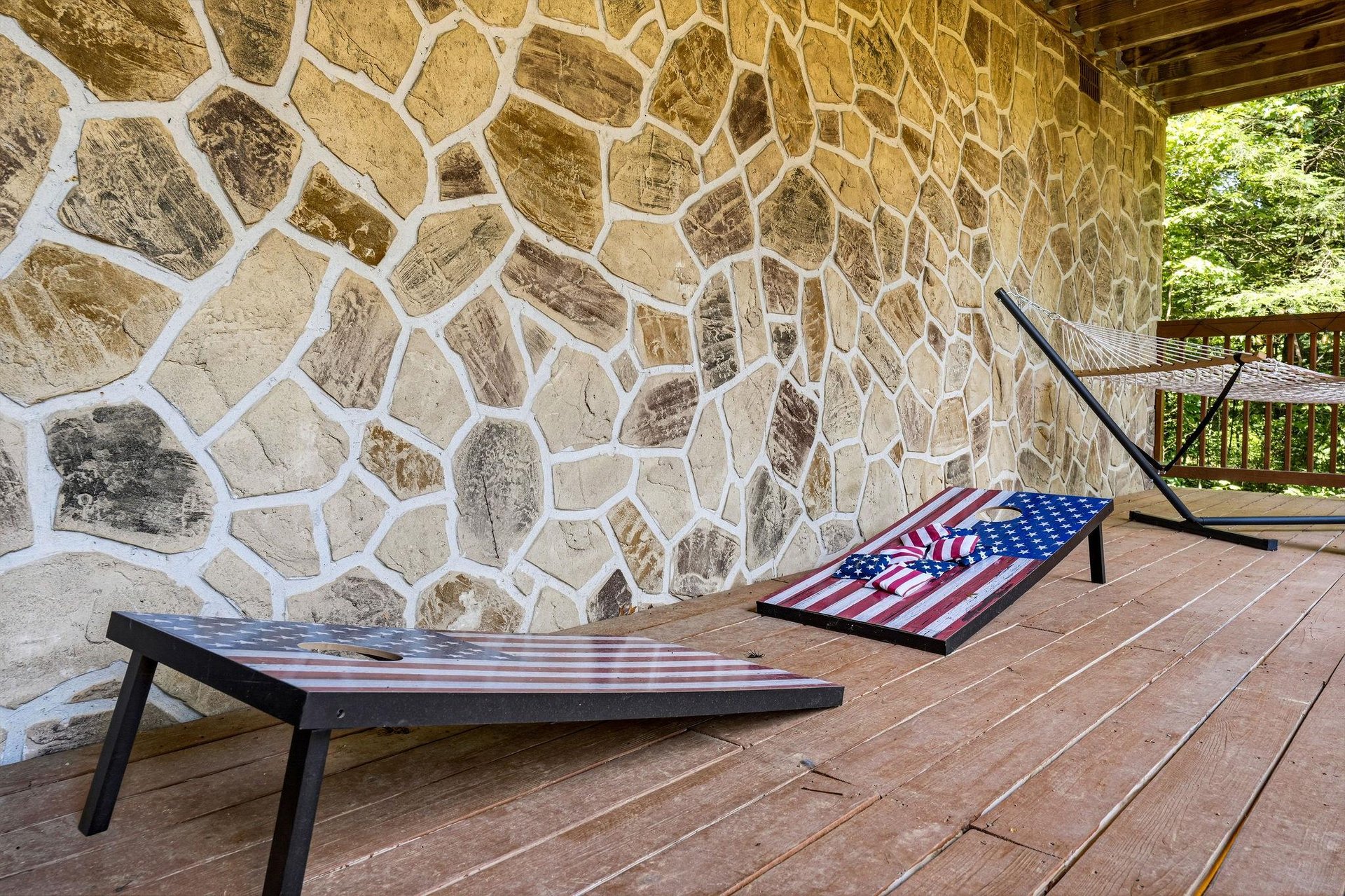 The deck features custom cornhole boards with patriotic designs, backed by rustic stone accents and surrounded by lush greenery—perfect for outdoor fun and friendly competition.