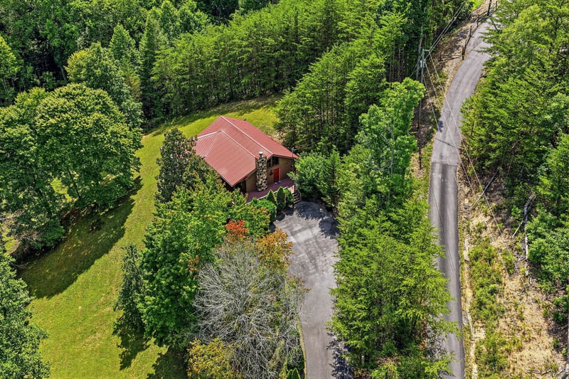 Aerial view of a secluded vacation rental nestled within lush forest, featuring a distinctive red roof and private driveway surrounded by mature trees and green lawn.