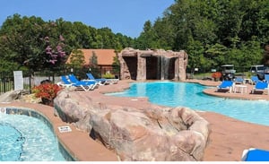 Resortstyle pool with waterfall feature surrounded by lush trees and comfortable lounge chairs under clear blue skies.