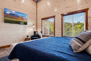 Wake up to bright natural light streaming through large windows, showcasing lush greenery beyond. The warm pine walls and plush bedding create a cozy retreat where you can truly unwind.