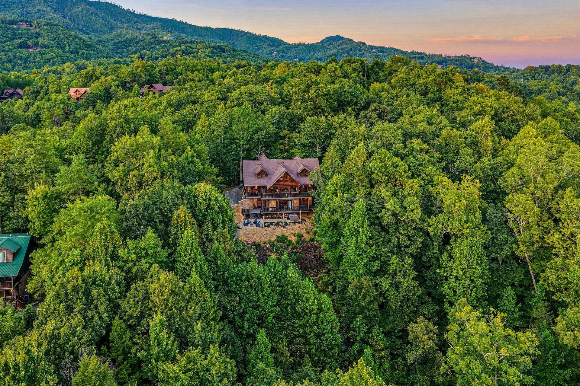 A stunning log cabin nestled in dense mountain forest with a vibrant sunset sky glowing above the surrounding peaks.
