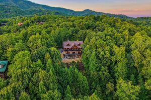 A stunning log cabin nestled in dense mountain forest with a vibrant sunset sky glowing above the surrounding peaks.