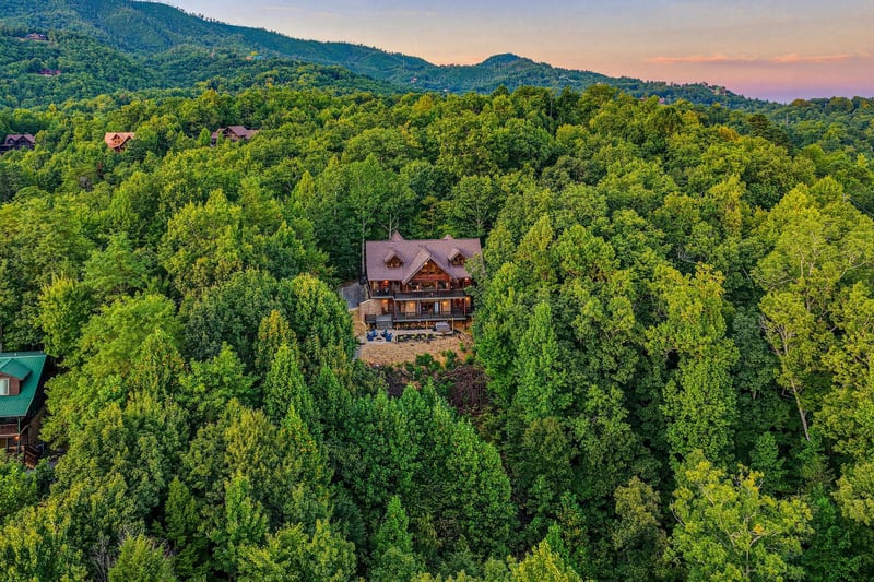 A stunning log cabin nestled in dense mountain forest with a vibrant sunset sky glowing above the surrounding peaks.