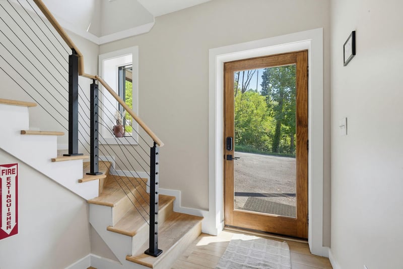 The entryway opens to a bright, airy staircase with modern cable railings and a warm wooden door framing views of lush greenery beyond.