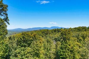 This elevated property offers sweeping views across lush forest and rolling ridges, with majestic mountains framing the horizon under a brilliant blue sky.