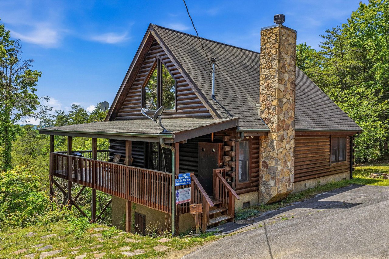 A charming log cabin with rustic stone chimney set among lush green trees in a peaceful mountain community with clear blue skies overhead.