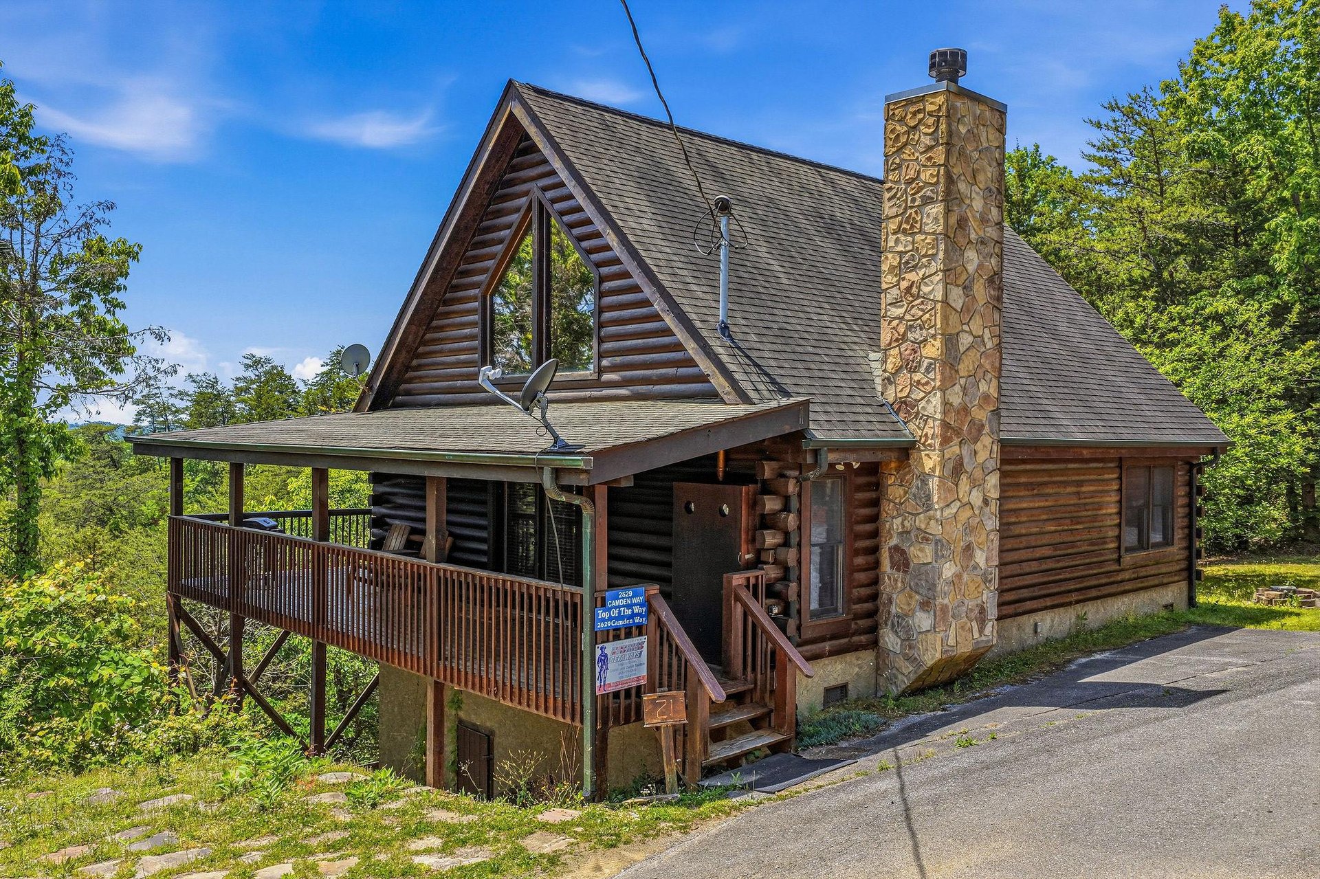 A charming log cabin with rustic stone chimney set among lush green trees in a peaceful mountain community with clear blue skies overhead.