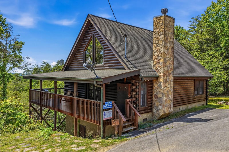 A charming log cabin with rustic stone chimney set among lush green trees in a peaceful mountain community with clear blue skies overhead.
