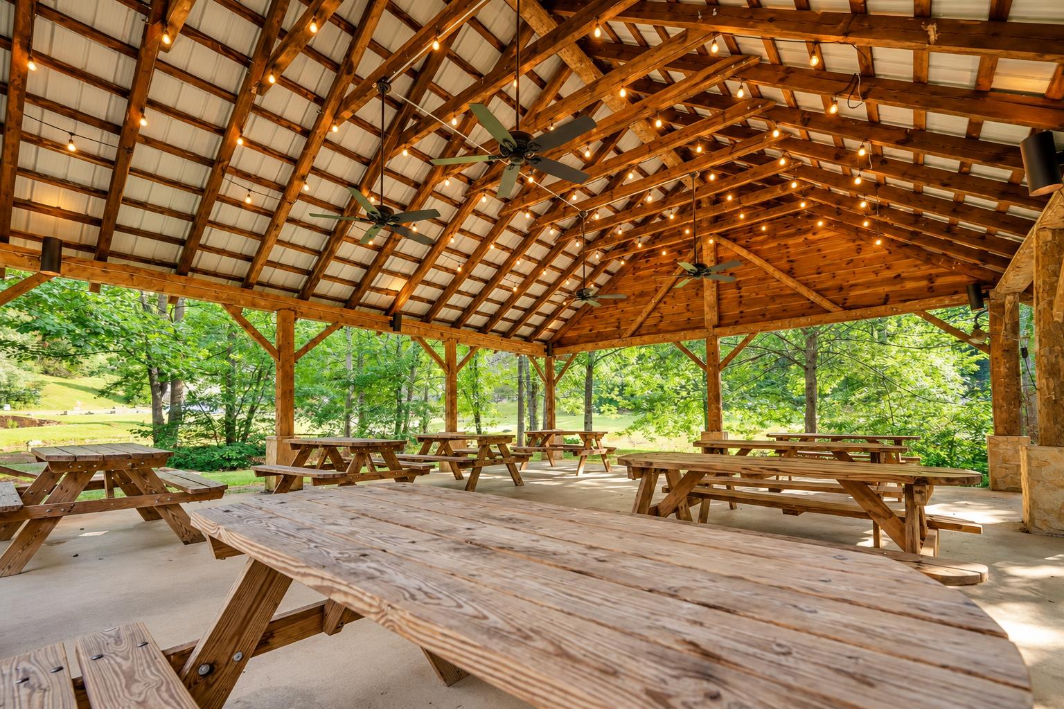 A spacious openair pavilion surrounded by lush greenery, featuring rustic picnic tables beneath a beautifully crafted vaulted wood ceiling strung with string lights—ideal for group gatherings and out