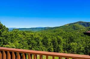 Expansive mountain views extend beyond a private balcony, with forested ridges layered beneath a bright blue summer sky creating a peaceful backdrop to the surrounding natural landscape.