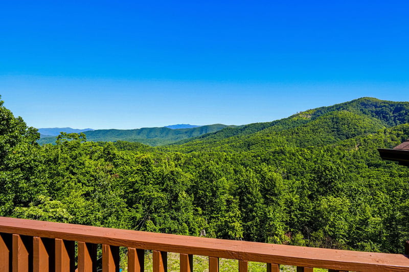 Expansive mountain views extend beyond a private balcony, with forested ridges layered beneath a bright blue summer sky creating a peaceful backdrop to the surrounding natural landscape.
