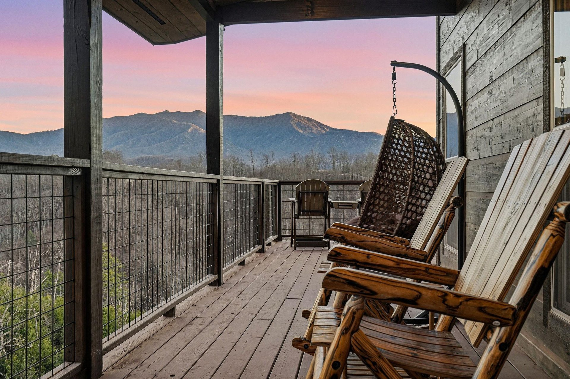 Covered deck with rustic rocking chairs and hanging swing chair offers stunning mountain views as the sky paints itself in sunset hues.