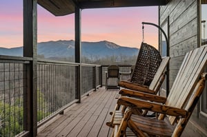 Covered deck with rustic rocking chairs and hanging swing chair offers stunning mountain views as the sky paints itself in sunset hues.