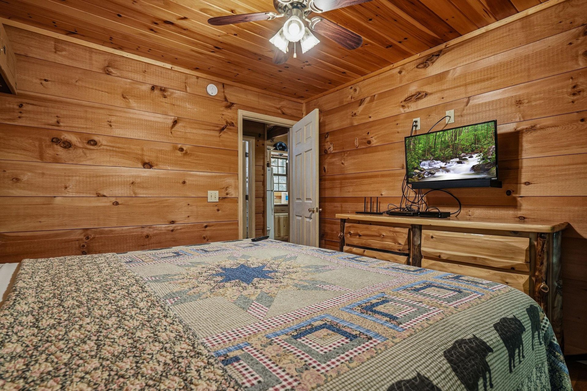 Unwind in your cozy cabin bedroom featuring warm pine walls, rustic log furnishings, and a ceiling fan for comfort. Enjoy your favorite shows on the flatscreen TV after exploring the outdoors.