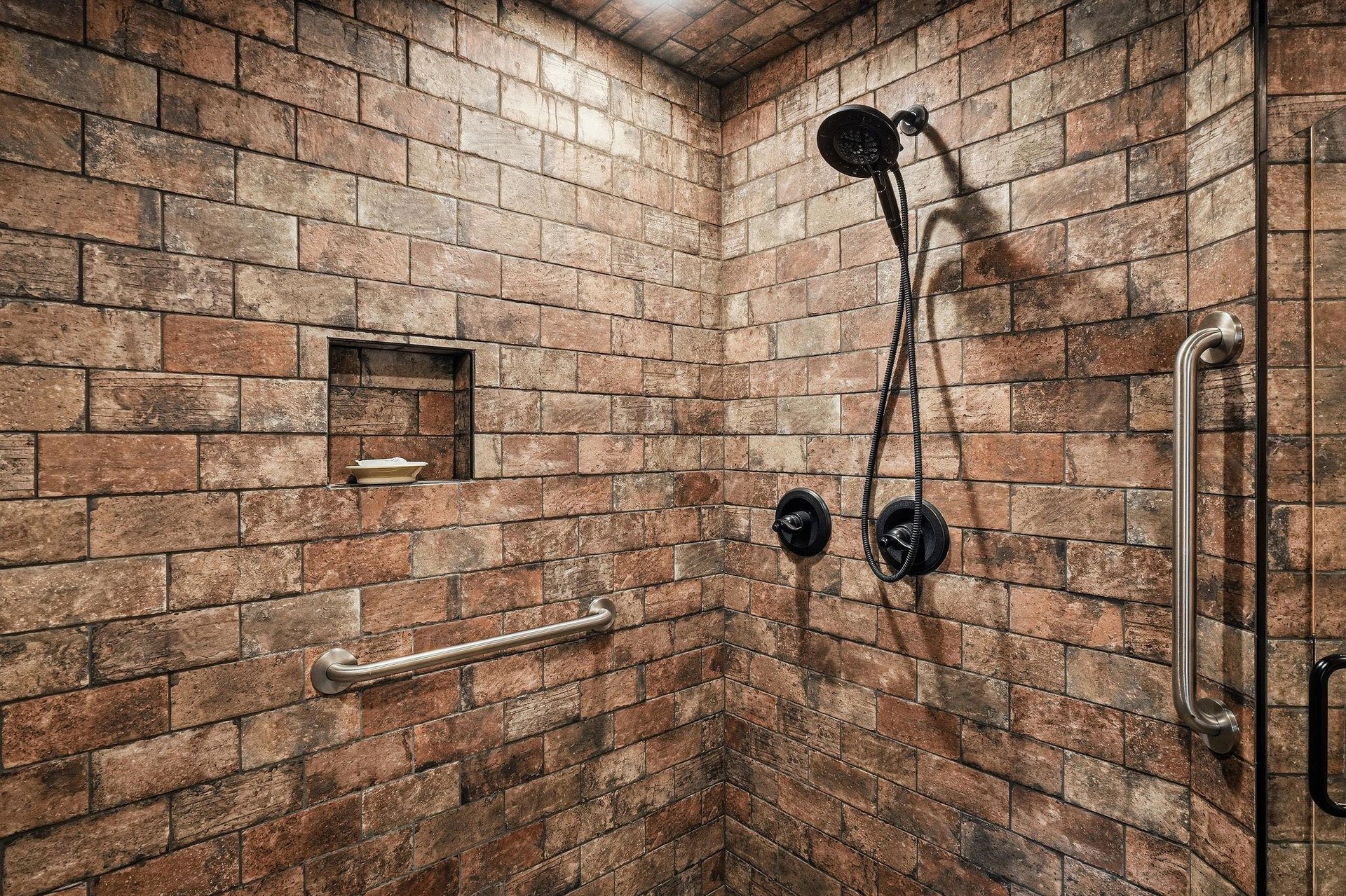 Spacious walkin shower with rustic tile, handheld and rain shower heads, builtin shelf, and safety grab bars for your comfort.