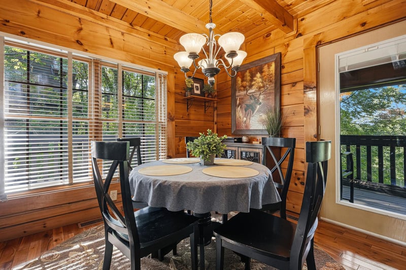 Your rustic dining nook features warm wood beams, elegant chandelier lighting, and peaceful forest views through large windows.