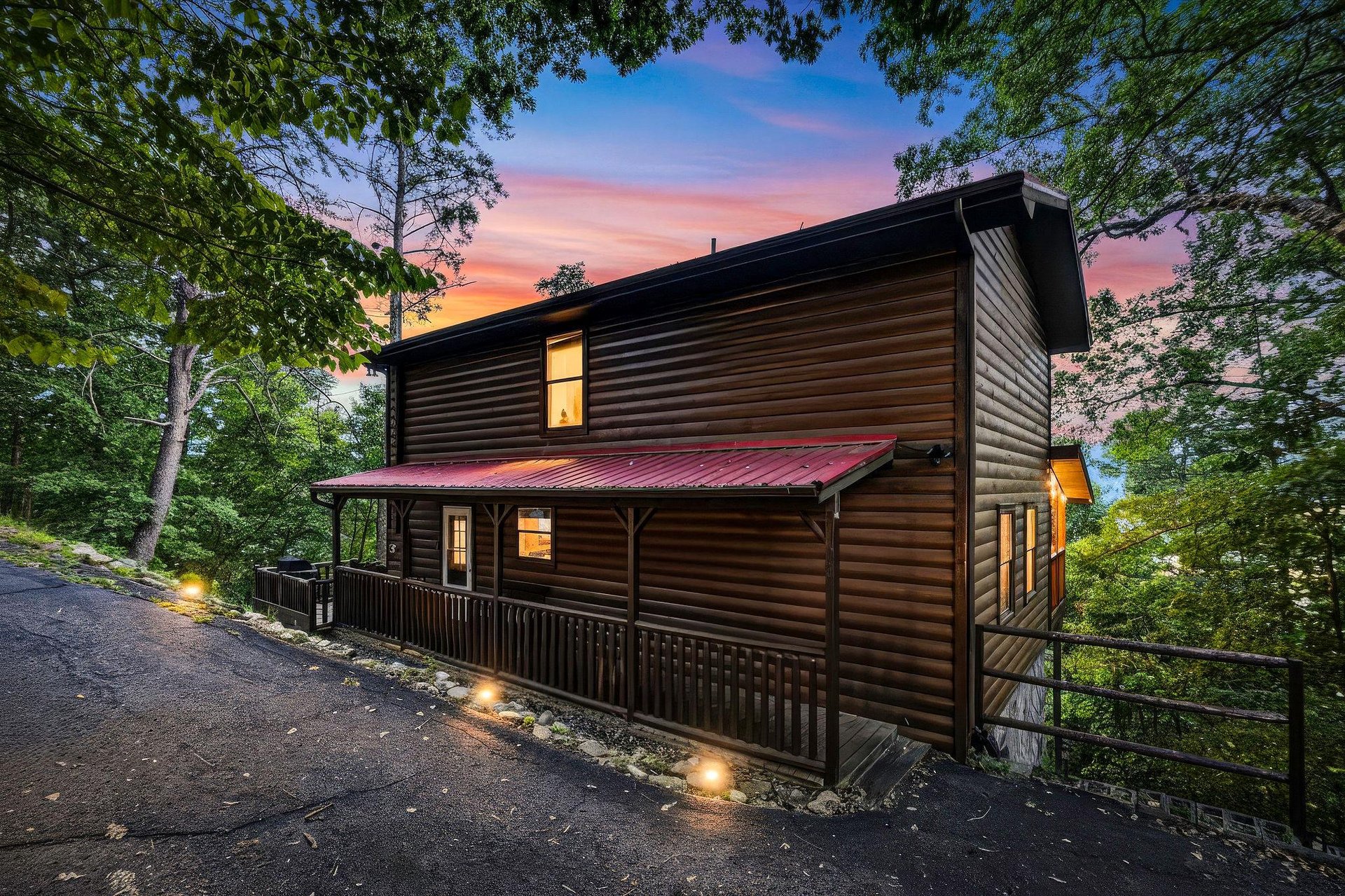 This property showcases a charming log cabin exterior nestled in a lush wooded setting, with warm evening lighting and a dramatic sunset sky overhead.