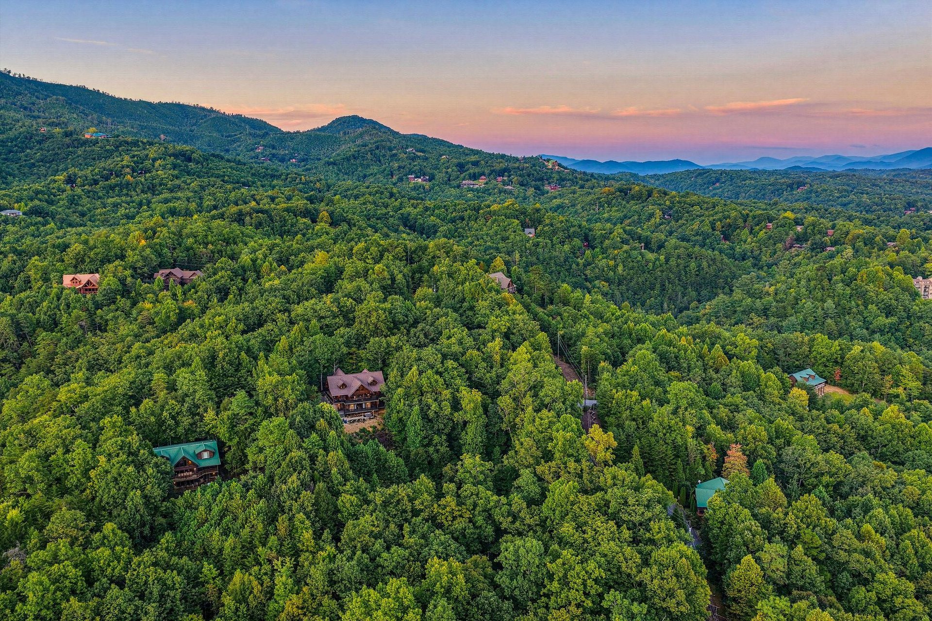 Aerial view of mountain cabins nestled among lush forested hills at sunset, showcasing the secluded natural setting of the property area.