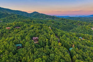 Aerial view of mountain cabins nestled among lush forested hills at sunset, showcasing the secluded natural setting of the property area.