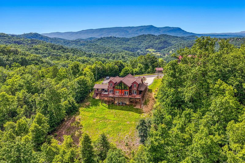 Aerial view of a red mountain lodge nestled among forested hills with sweeping mountain vistas. The property sits on a hilltop clearing surrounded by natural landscape and wilderness.