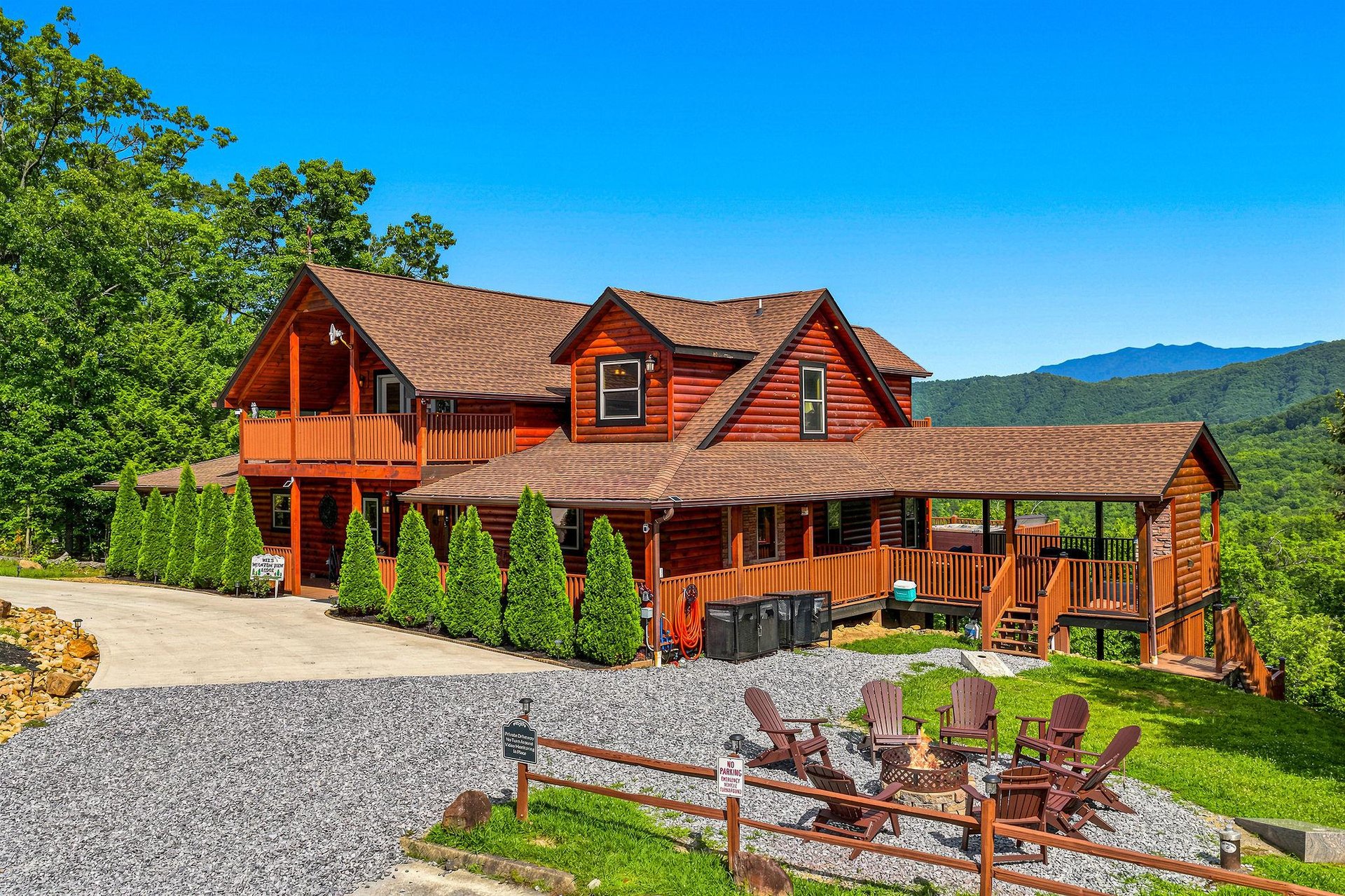 A spacious mountain cabin featuring natural log construction, expansive wraparound decks, and stunning mountain backdrop. The property includes a gravel fire pit area with seating and hot tub access.