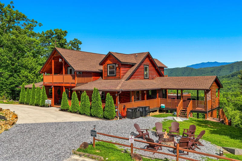 A spacious mountain cabin featuring natural log construction, expansive wraparound decks, and stunning mountain backdrop. The property includes a gravel fire pit area with seating and hot tub access.