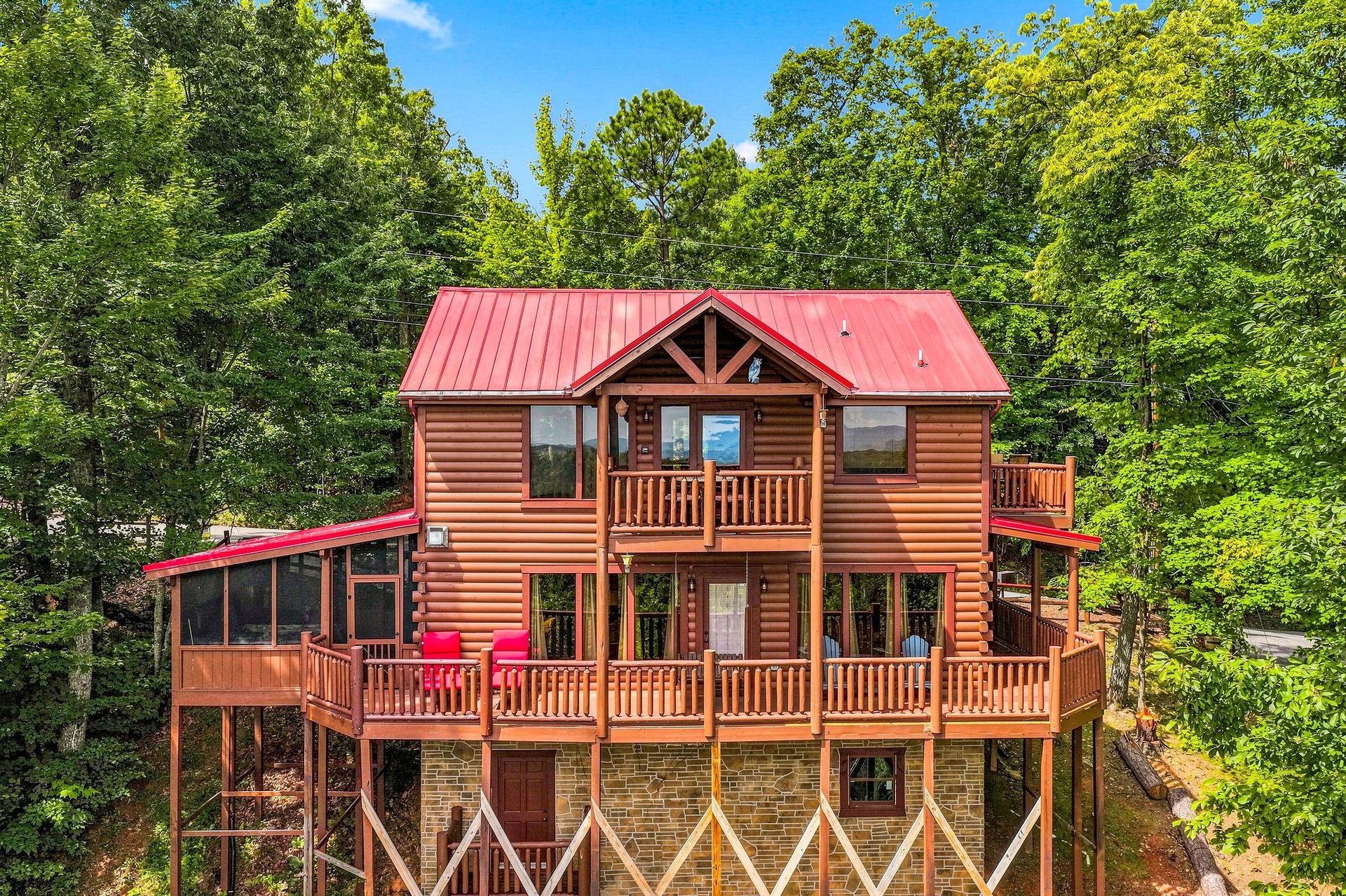 A charming log cabin nestled among lush forest, complete with spacious multilevel decks and bright red accents under a clear blue sky.