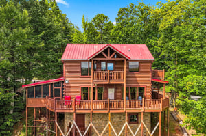 A charming log cabin nestled among lush forest, complete with spacious multilevel decks and bright red accents under a clear blue sky.