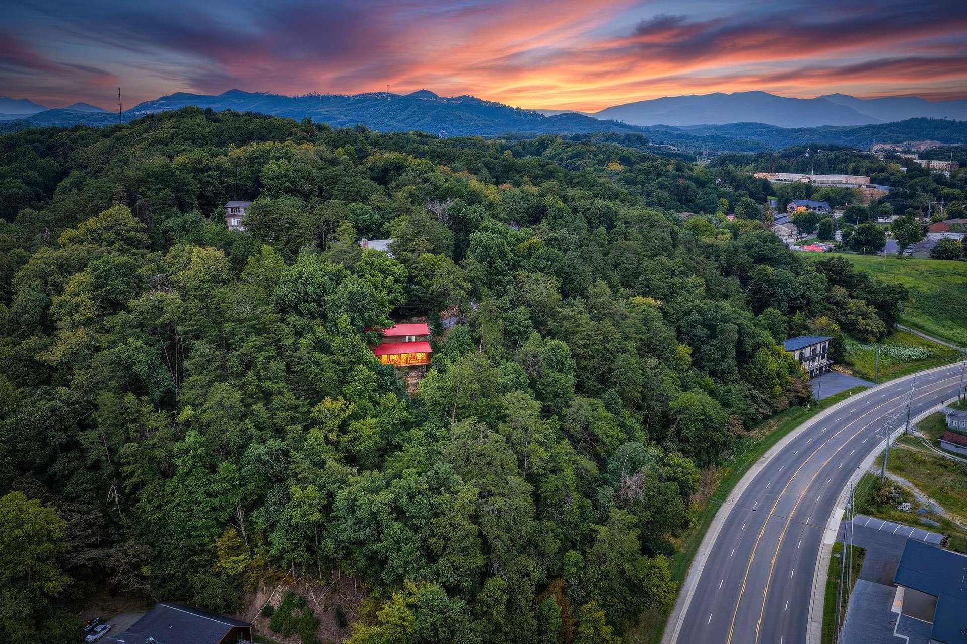 A stunning aerial view captures the property nestled in lush forested hills with dramatic mountain ridges and a vibrant sunset sky painting the horizon in coral and purple hues.