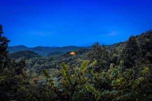 The property sits nestled in the surrounding mountainous terrain, with forested hillsides and a distant structure visible against the evening sky.