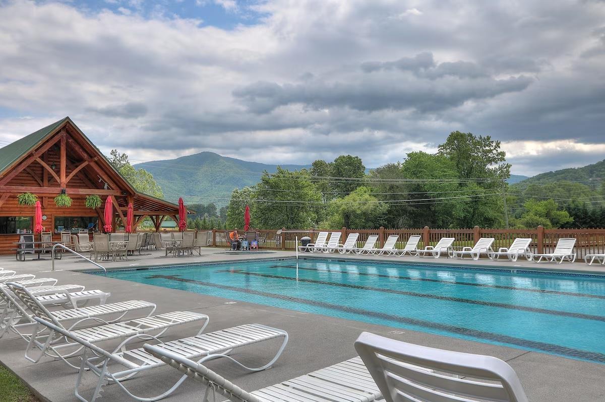Resort pool area with mountain backdrop and rustic lodgestyle building featuring outdoor seating and scenic wilderness views.