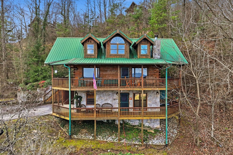 Charming mountain cabin with distinctive green metal roof and wraparound decks nestled in wooded hillside setting.
