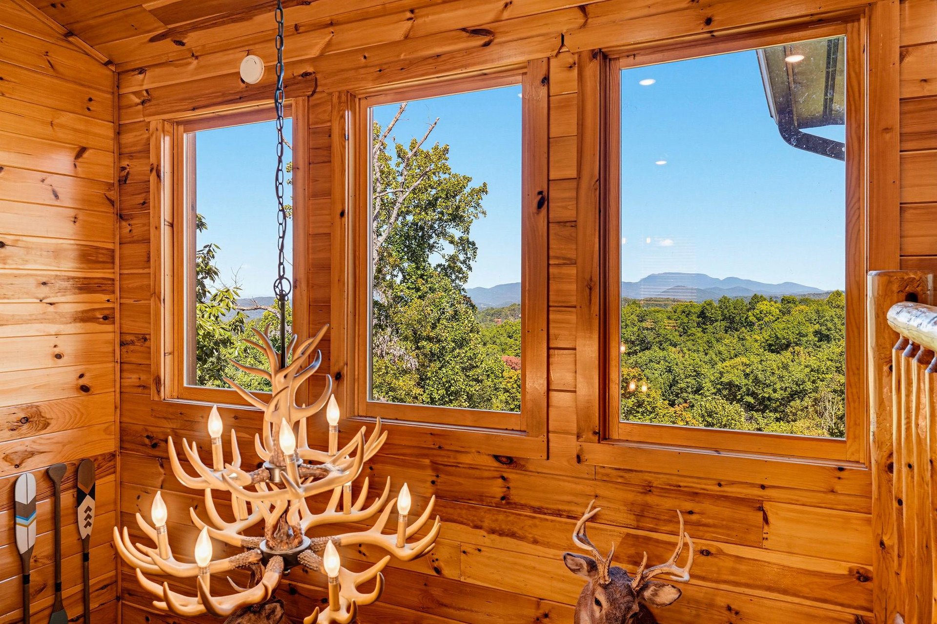 Sunlight streams through expansive windows framing sweeping mountain vistas and lush forests, while rustic wood paneling and an antler chandelier create a cozy, lodgelike retreat youll love.