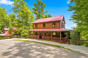 A charming log cabin with a red metal roof and wraparound porch sits nestled among tall trees. The spacious driveway offers easy parking.