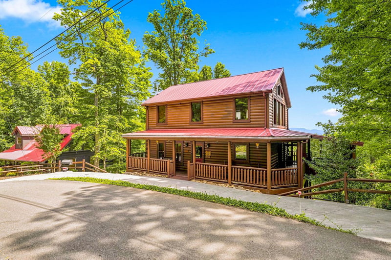 A charming log cabin with a red metal roof and wraparound porch sits nestled among tall trees. The spacious driveway offers easy parking.