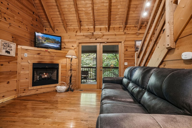 Relax in your cozy cabin living room with vaulted wood ceilings, a warm fireplace, and balcony doors opening to peaceful forest views. Sink into the plush leather sofa and unwind.