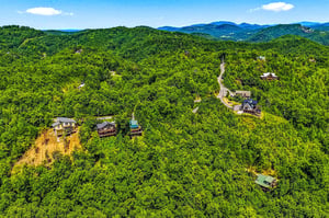 A scenic mountain community nestled in lush forest, with winding roads connecting charming cabinstyle homes beneath distant blue peaks and summer skies.