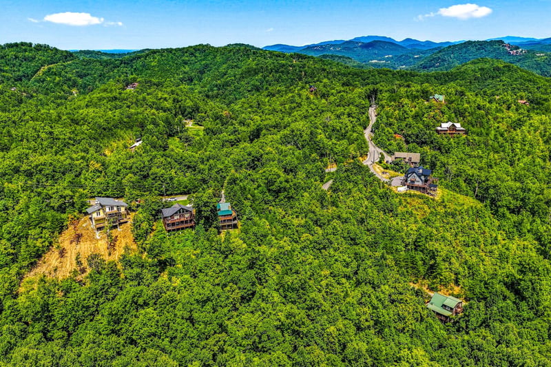 A scenic mountain community nestled in lush forest, with winding roads connecting charming cabinstyle homes beneath distant blue peaks and summer skies.