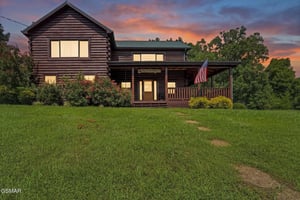 A charming logstyle cabin featuring a welcoming front porch, surrounded by lush green lawn and mature trees beneath a stunning sunset sky.