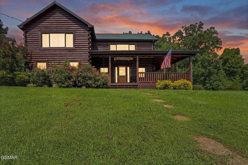 A charming logstyle cabin featuring a welcoming front porch, surrounded by lush green lawn and mature trees beneath a stunning sunset sky.