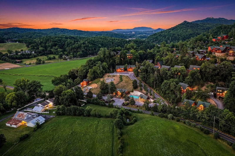 Aerial view captures the property nestled among rolling mountains and lush forests, with warm cabin lights glowing against a stunning sunset sky over the surrounding valley.
