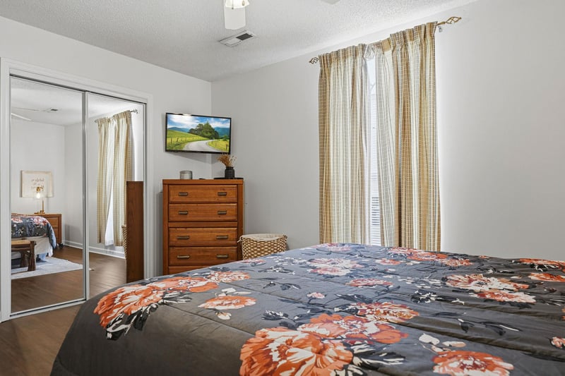 Unwind in this bright, cozy bedroom with a plush bed, warm wood furnishings, flatscreen TV, and natural light streaming through elegant curtains—your peaceful retreat after a day of exploring.