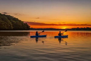 Two kayakers paddle across a serene lake as the sun sets, painting the water with vibrant golden and orange hues—a perfect backdrop for nearby water activities.