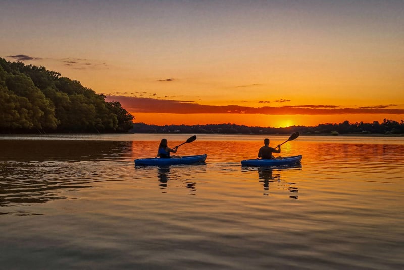 Two kayakers paddle across a serene lake as the sun sets, painting the water with vibrant golden and orange hues—a perfect backdrop for nearby water activities.